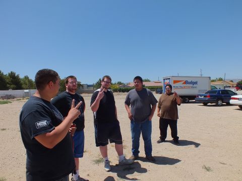 2003 Ford Crown Victoria - group of men converse on a sandy lot, one in an NOS shirt, a Budget truck parked behind