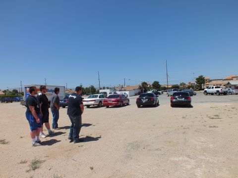 2003 Ford Crown Victoria - cluster of people stand on a gravel lot beside parked sedans, pickups and a rental truck