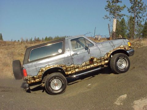 1989 Ford Bronco - Passenger-side view of a lifted gray full-size Ford Bronco with black removable hardtop, posed on a dirt embankment with the front wheel li…