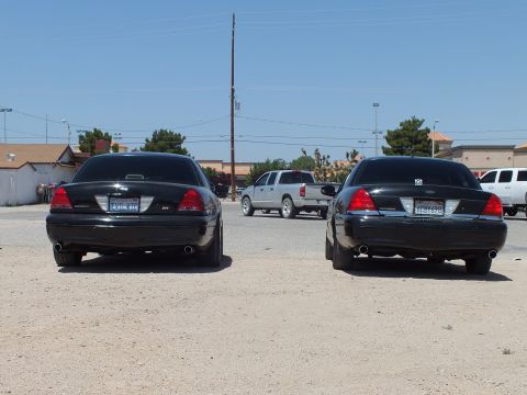 2003 Ford Crown Victoria - pair of black full-size sedans, one badged SVT, sit on gravel with a pickup passing behind