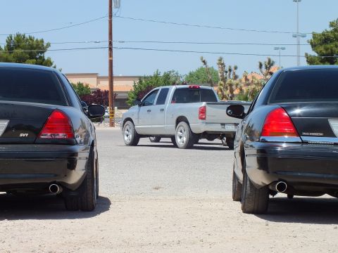 2003 Ford Crown Victoria - Two black sedans with SVT badging frame a silver Dodge Ram crew cab across the gravel lot