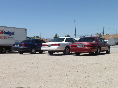 2003 Ford Crown Victoria - Three American sedans staged on a dusty lot next to a Budget moving truck under blue sky