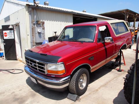 1994 Ford Bronco - A red 1994 Ford Bronco is parked on jack stands outside a building, undergoing maintenance.
