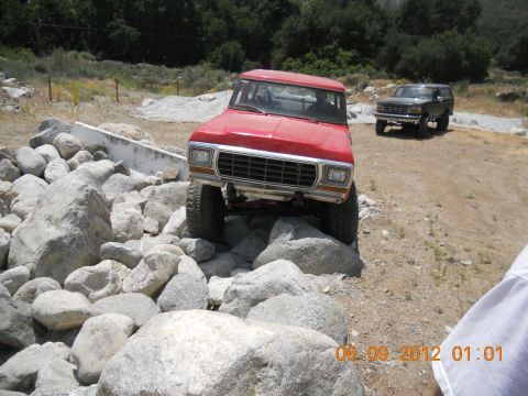1986 Ford Bronco - Two Ford Broncos are seen off-roading on a rocky trail. The front vehicle is red, the rear is dark.