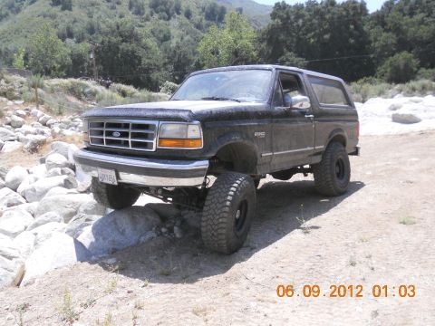1986 Ford Bronco - This lifted black Ford Bronco is shown off-roading on a sunny day, with rocks and trees in the background.