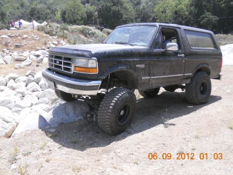1986 Ford Bronco - A lifted black Ford Bronco navigates rocky terrain on a sunny day.