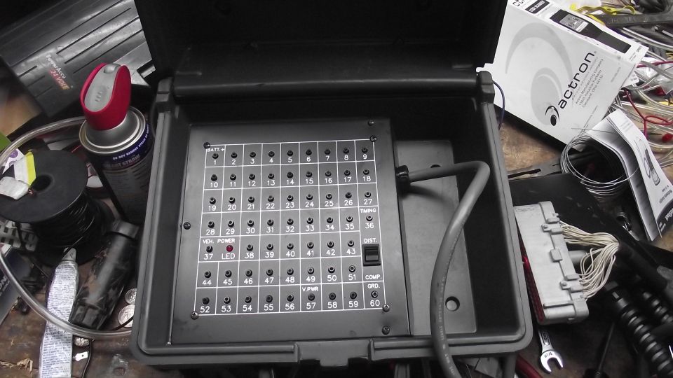 Open black box with a grid of numbered fuse slots, some labeled. Wires and a manual are nearby