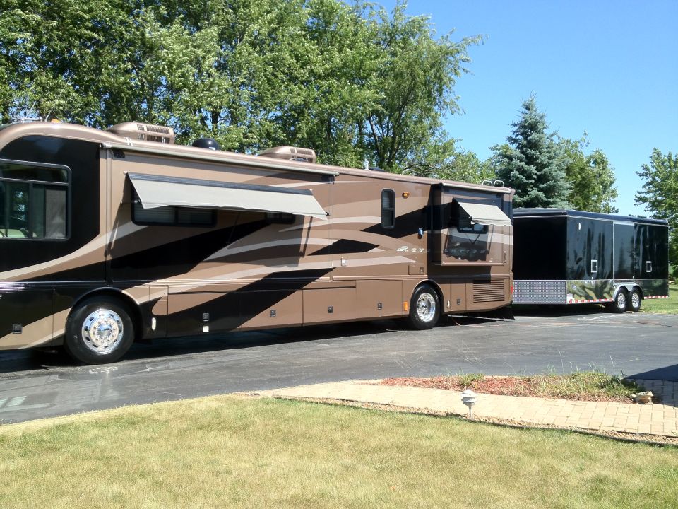 Motorhome towing a black cargo trailer. Both vehicles are parked on a paved surface with grass and trees in the background
