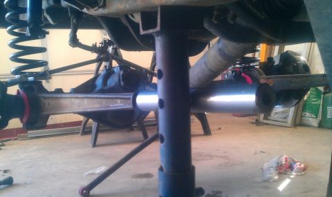 1987 Ford Bronco - The underside of a vehicle is shown, with the rear axle assembly supported by a jack stand. A silver metal tube is positioned horizontally…