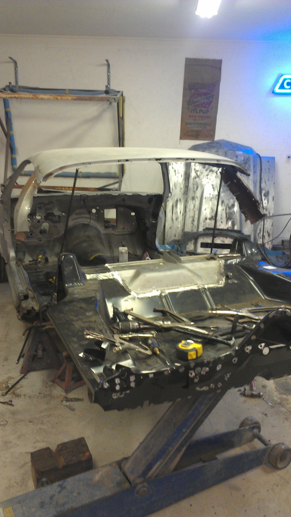 Car body on lift, stripped down for restoration. Tools and parts are visible around the chassis