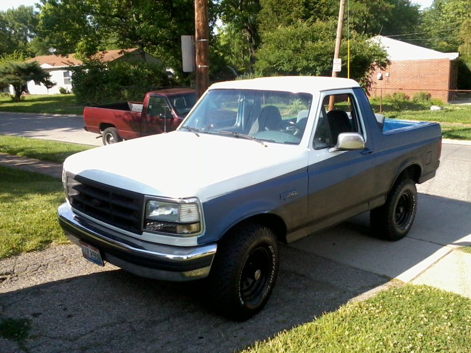 Two-tone white and blue Ford Bronco with top removed, black wheels, parked in a driveway, front three-quarter view