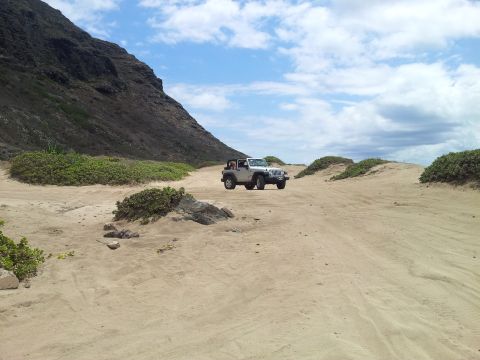 1990 Chevrolet Suburban - A silver Jeep Wrangler navigates a sandy terrain near a coastal mountain under a cloudy sky.