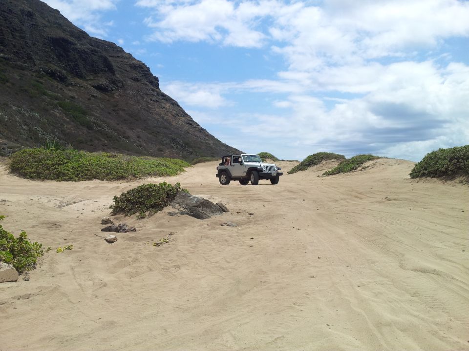 Silver Jeep Wrangler driving on a sandy beach with a large mountain in the background