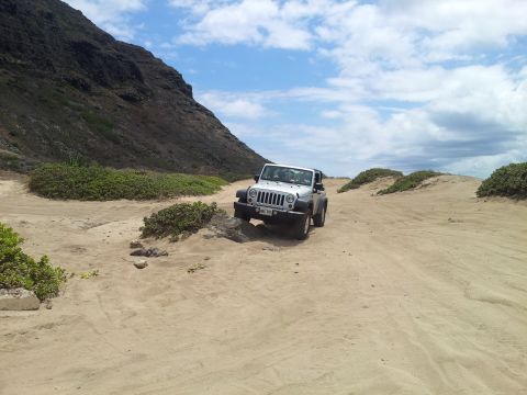 1990 Chevrolet Suburban - Jeep Wrangler navigating a sandy path near coastal hills under a partly cloudy sky.