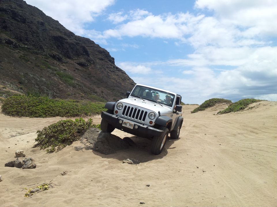 White Jeep Wrangler driving off-road on sand and rocks, with a mountain and cloudy sky