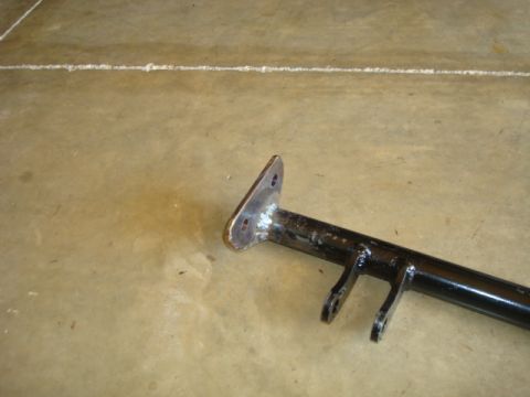 1995 Ford Bronco - black-painted fabricated steel suspension or chassis arm with a flat mounting flange and clevis tabs, lying on a concrete floor.