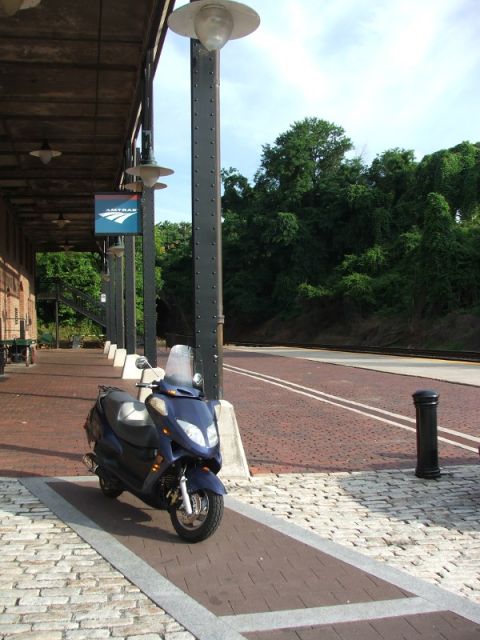 2008 Yamaha Zuma - A blue scooter is parked on a train station platform next to the tracks. An Amtrak sign is visible in the background.