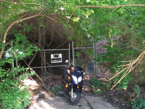 2008 Yamaha Zuma - A dark scooter sits before a fenced-off, overgrown area with graffiti on the wall.
