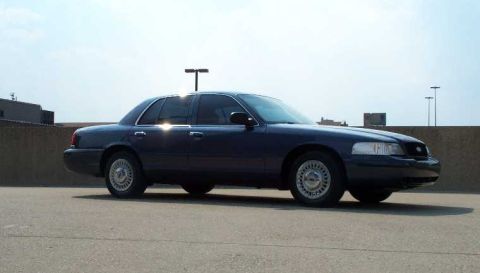 1998 Ford Crown Victoria - A dark blue 1998 Ford Crown Victoria sedan is parked on asphalt under a bright sky.