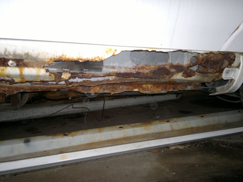 Close-up of a white vehicle's rocker panel showing extensive rust and holes. The panel is heavily corroded, with layers of rust flaking off