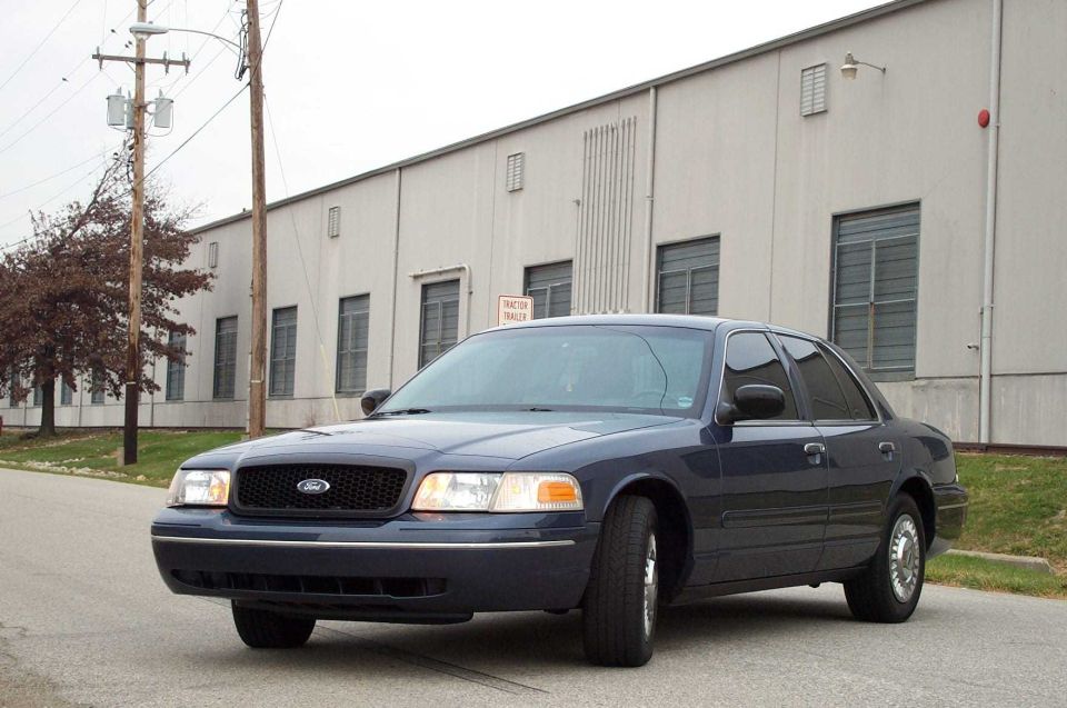 Dark blue Ford Crown Victoria sedan, front 3/4 view, parked by gray industrial building.