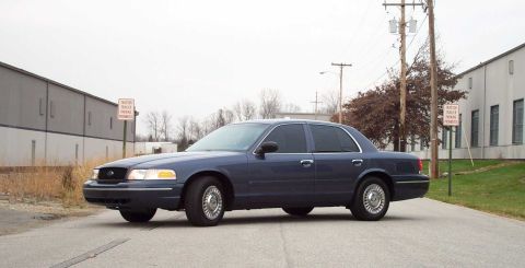 1998 Ford Crown Victoria - A blue sedan is parked on a street in front of industrial buildings under an overcast sky.