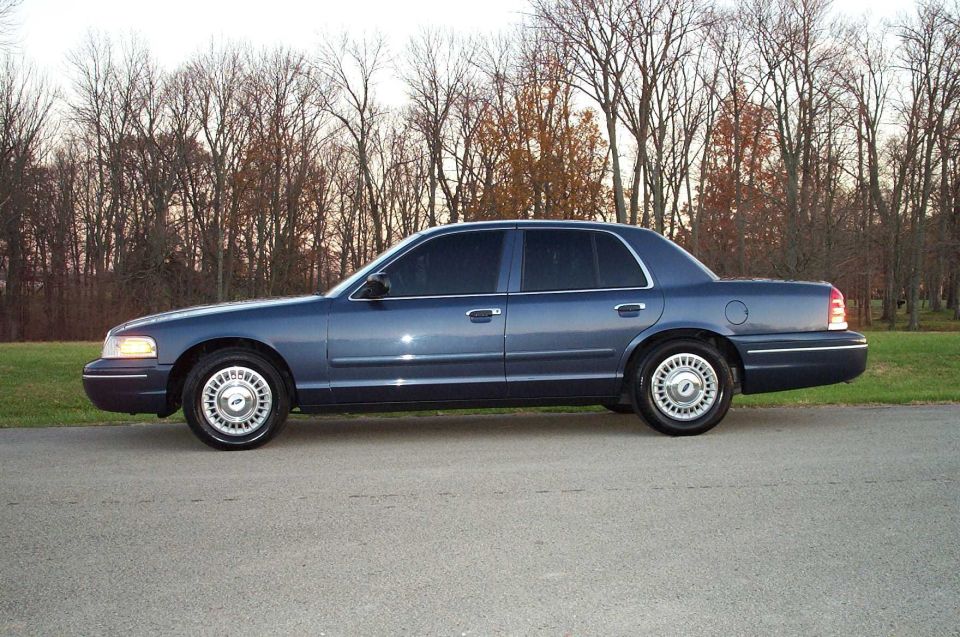 Dark blue Ford Crown Victoria sedan in side profile on pavement with trees behind.