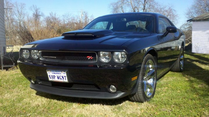 Black Dodge Challenger parked on grass, front quarter view. License plate says KOP MGNT