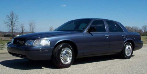 1998 Ford Crown Victoria - This blue sedan is parked outdoors under a clear blue sky with trees in the distance.