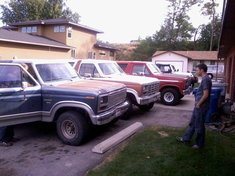 Several Ford Broncos and F-Series trucks parked in a driveway. One blue Bronco is in the foreground with its door open