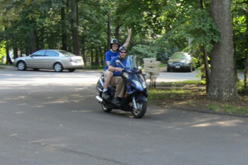 2008 Yamaha Zuma - Two people ride a blue scooter down a paved road with trees and cars visible.