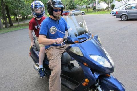 2008 Yamaha Zuma - A man and a child ride on a blue scooter, both wearing helmets and sunglasses.
