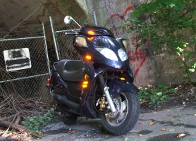 2008 Yamaha Zuma - A dark blue scooter is parked outdoors near a fence with graffiti on a concrete structure.
