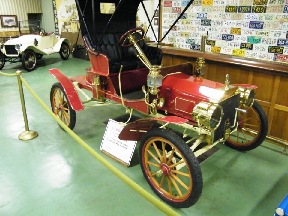 red touring car with black top, wooden spoke wheels, and brass accents, parked indoors near a white car