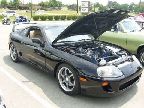 2007 Ford Mustang - A black Toyota Supra is parked with its hood open, revealing the engine and turbocharger. The car is seen from a three-quarter front view.