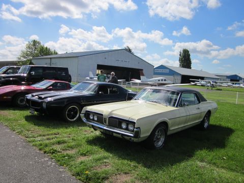 1991 Ford LTD Crown Victoria - A line of vintage cars are parked on the grass in front of airplane hangars under a cloudy sky.