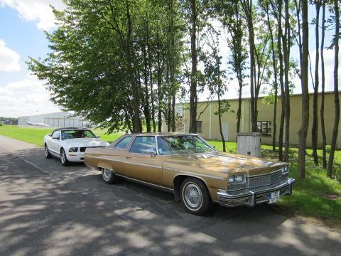 1991 Ford LTD Crown Victoria - A gold 1976 Buick LeSabre coupe is parked next to a white convertible, with trees and a building in the background.