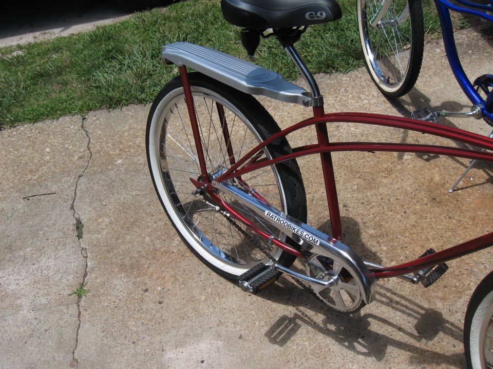 Rear section of a red cruiser bicycle with whitewall tire, chrome fender, and chain guard. Another blue bicycle is visible behind it