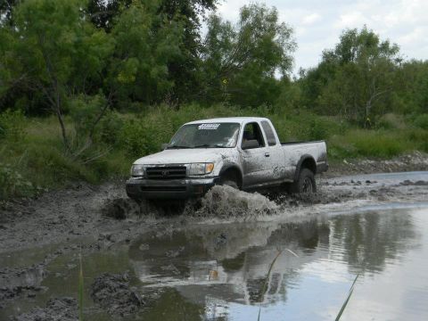 1989 Ford Bronco - White Toyota Tacoma navigates a muddy off-road path, kicking up water.