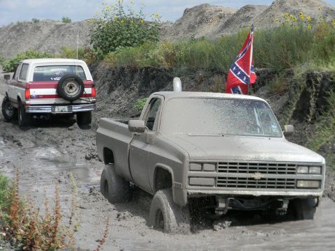 1989 Ford Bronco - Two off-road vehicles navigate a muddy terrain, one with a Confederate flag flying.