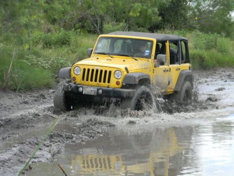 1989 Ford Bronco - Yellow Jeep Wrangler navigates a muddy trail, splashing water as it goes.