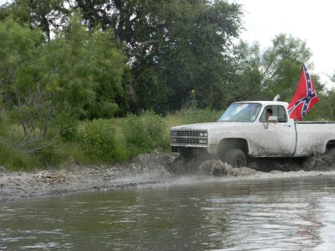 1989 Ford Bronco - A white pickup truck is driving through a muddy, watery area with trees in the background and a Confederate flag flying from the bed.