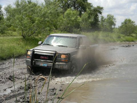 1989 Ford Bronco - A GMC pickup truck navigates a muddy off-road path, kicking up water and dirt.