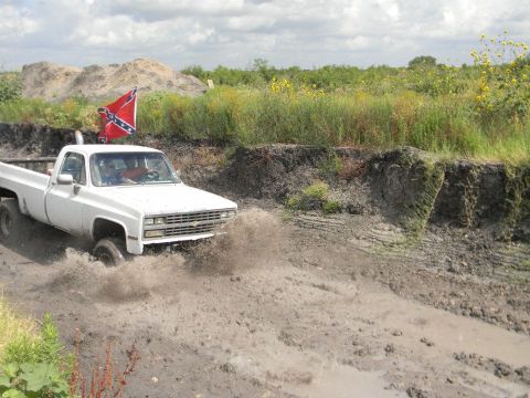 1989 Ford Bronco - A white pickup truck navigates a muddy terrain with a Confederate flag attached to its antenna.