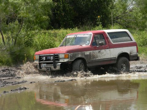 1989 Ford Bronco - This Ford Bronco is navigating a muddy terrain, kicking up water and mud as it goes.