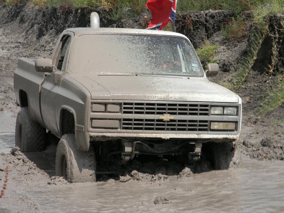 Mud-covered pickup truck stuck in mud, front view. Grille indicates 1980s Chevrolet