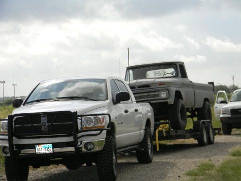 1989 Ford Bronco - A white pickup truck tows a grey pickup truck on a trailer, with another vehicle visible in the background.