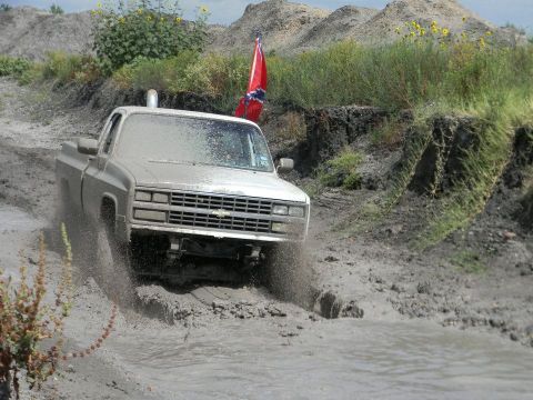 1989 Ford Bronco - This pickup truck is navigating a muddy off-road course, with a rebel flag flying from a pole on its side.
