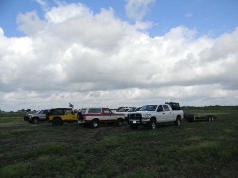 1989 Ford Bronco - Several SUVs and trucks are parked in a wide, grassy area under a cloudy sky.