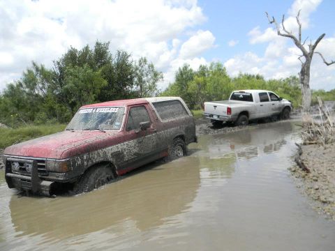 1989 Ford Bronco - Muddy 1989 Ford Bronco navigates a waterlogged trail, followed by a white pickup truck.